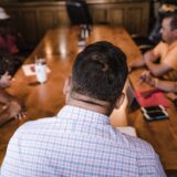 Grup de oameni de vârste diferite care discută în jurul unei mese, la o întâlnire a unei asociații locale